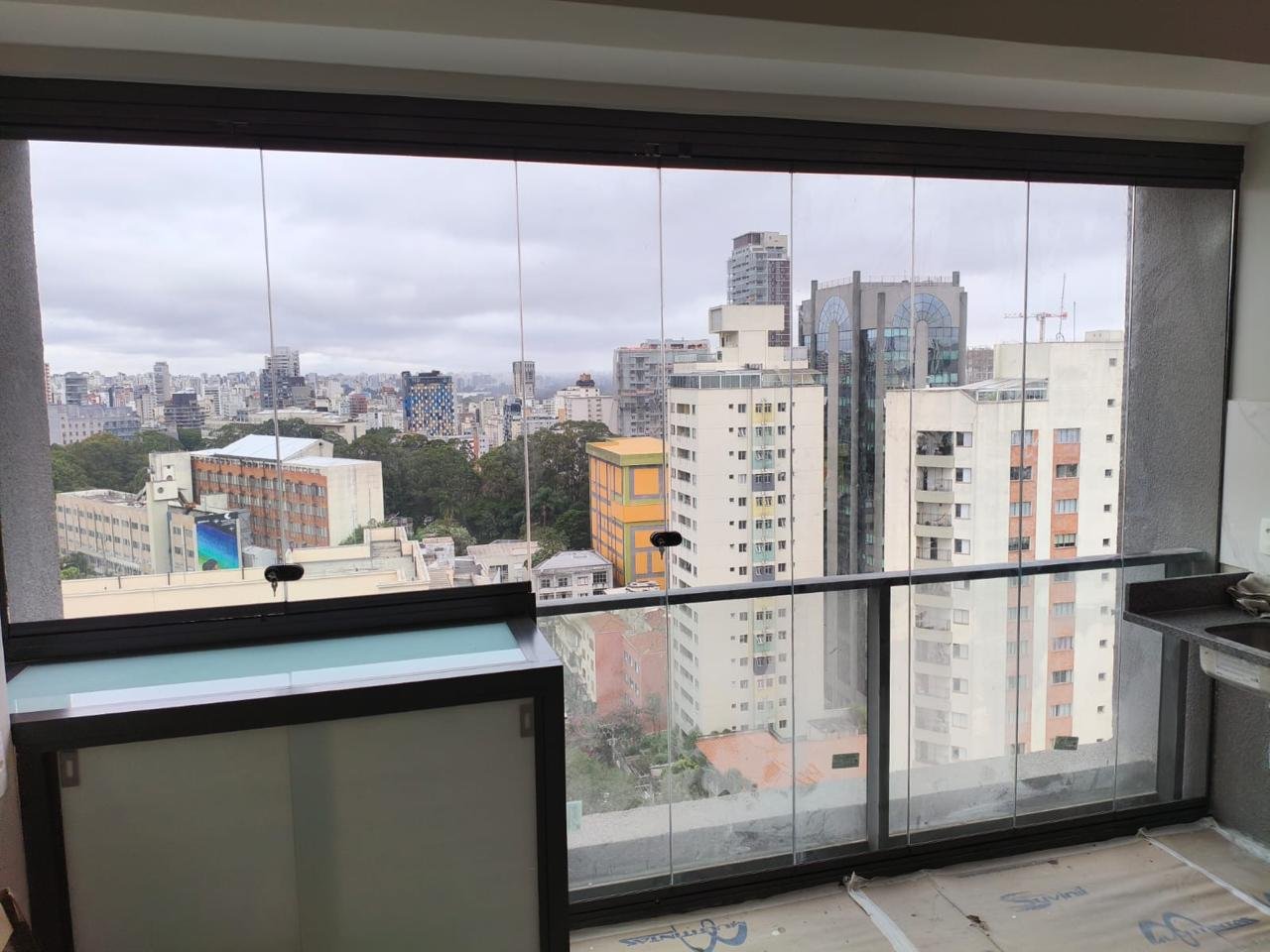 Sacada envidraçada com perfis pretos, bancada à esquerda e pia à direita, com vista para prédios e árvores
